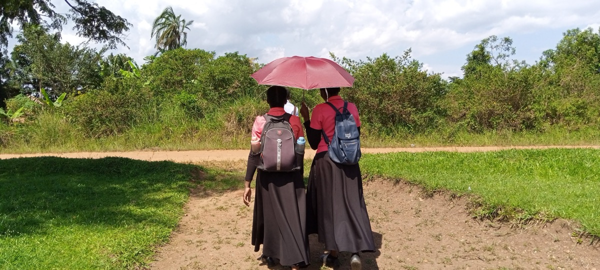 Two Labwor Missions women walking together under a shared umbrella along a village path in Uganda